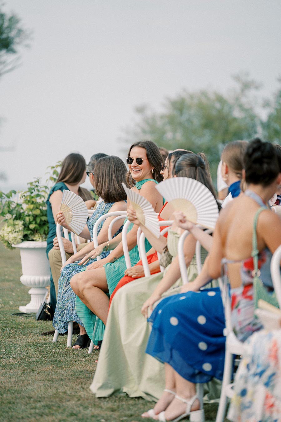 Guests in colorful dresses sitting outdoors, holding fans, and smiling at a summer event.