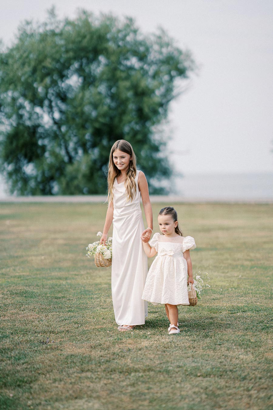 Two young girls in white dresses walking across a grassy field, each holding a small wicker basket with flowers, under a clear sky with trees in the background.