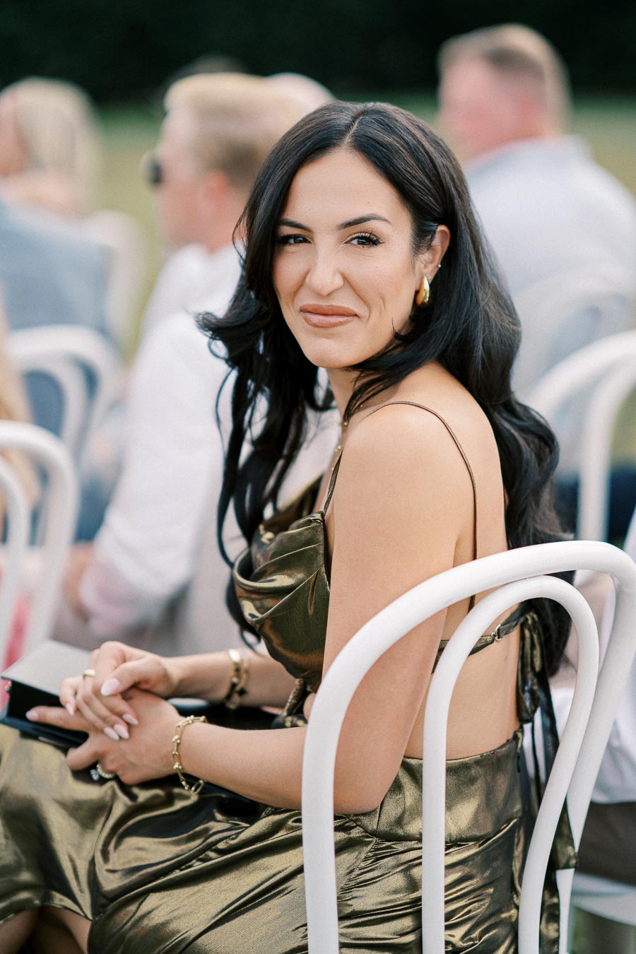 A woman in a stylish metallic dress sitting on a white chair at an outdoor event, smiling at the camera.