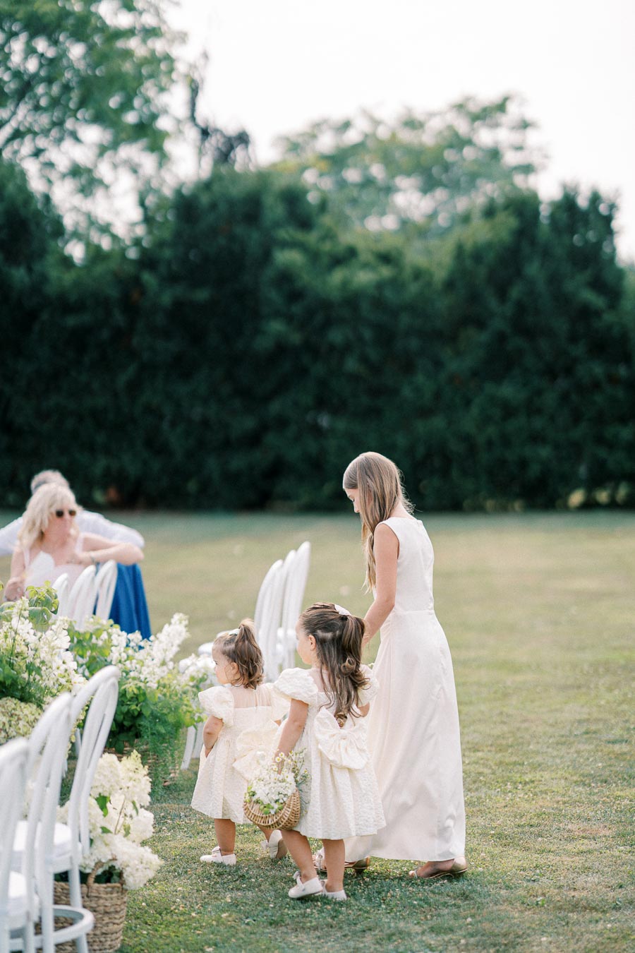 Three young girls in white dresses walking on a grassy area, surrounded by lush greenery, during an outdoor event with white chairs and floral decorations.