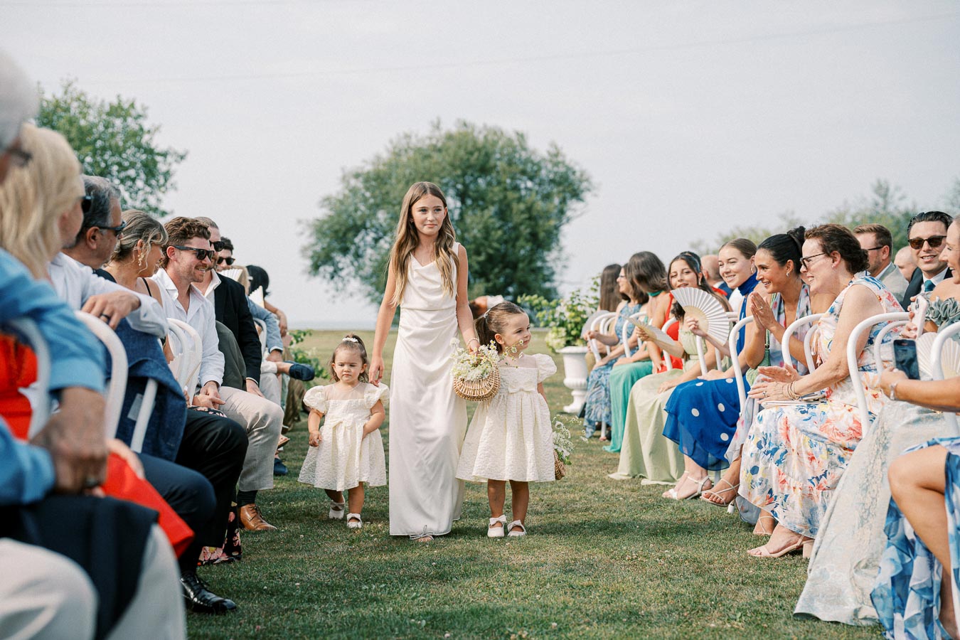 Outdoor wedding ceremony with a young girl and two children walking down the aisle, surrounded by seated guests in festive attire, beneath a clear sky and lush greenery.