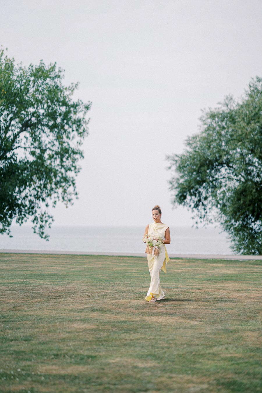 A woman in a yellow dress holding a bouquet walks on a grassy field with trees and a lake in the background.