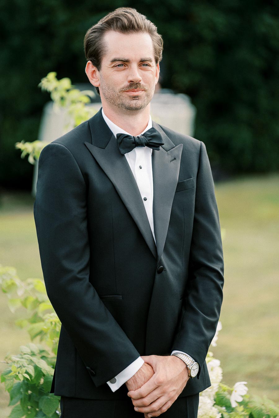 A man in a formal black tuxedo with a bow tie stands outdoors, surrounded by greenery, looking confidently at the camera.