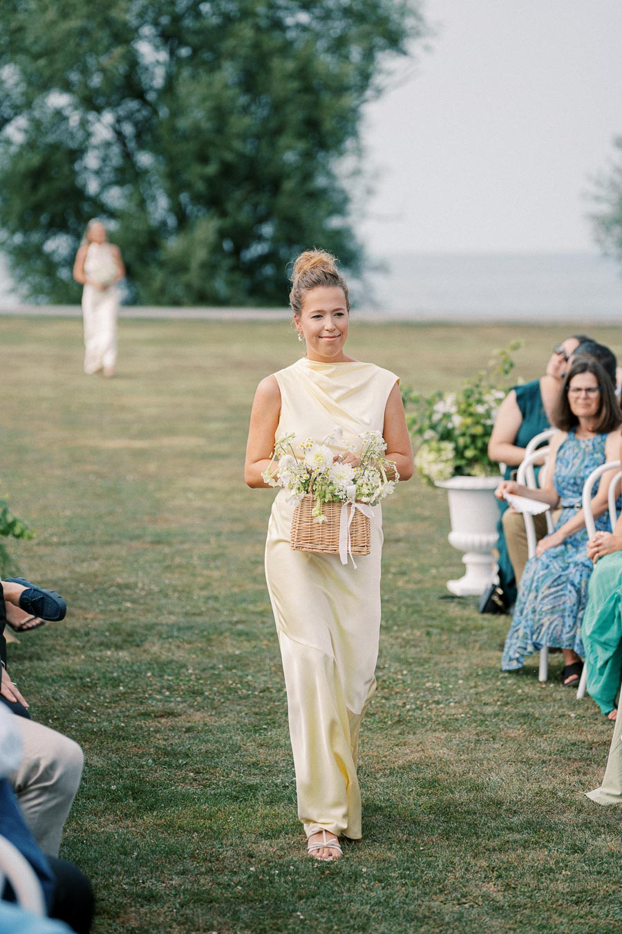 Bridesmaid in a pale yellow dress walking down a grassy aisle at an outdoor wedding ceremony, holding a wicker basket filled with flowers, surrounded by seated guests.