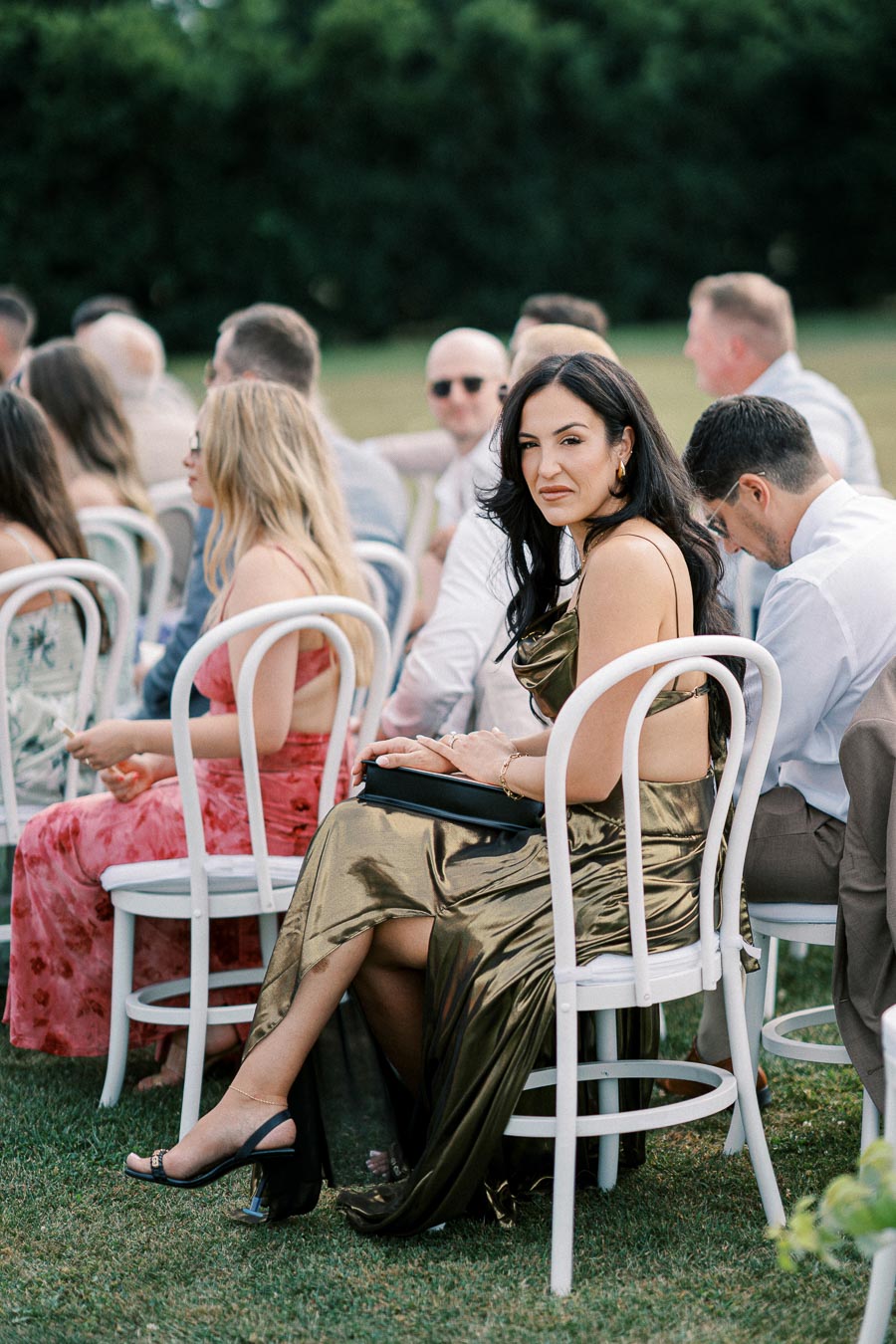 A group of elegantly dressed guests seated outdoors at a wedding ceremony, featuring a woman in the foreground wearing a metallic gold dress and holding a clutch.