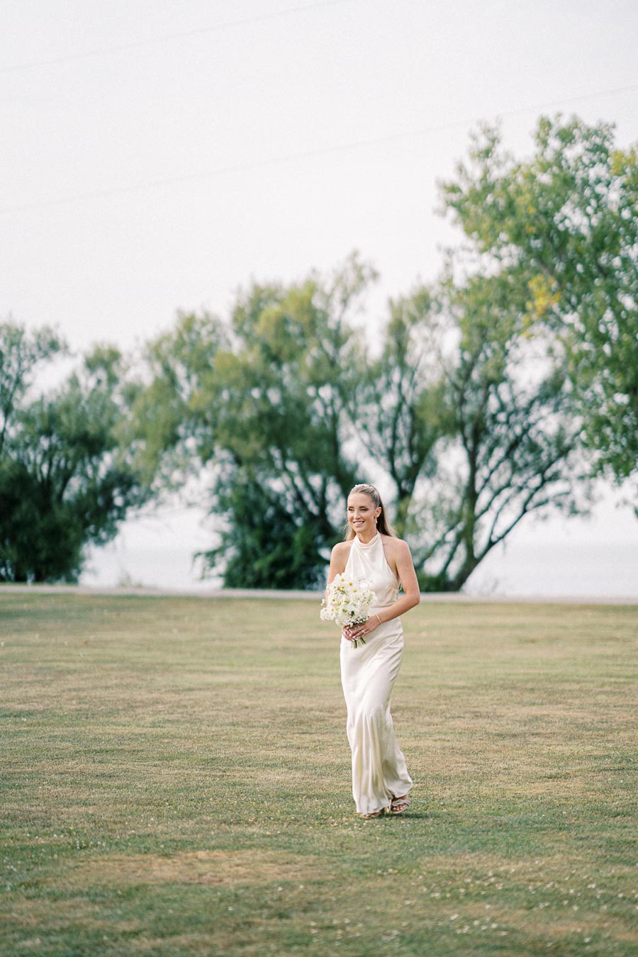 A bride in an elegant white dress walking on a grassy field holding a bouquet, with trees and a serene background during an outdoor wedding ceremony.