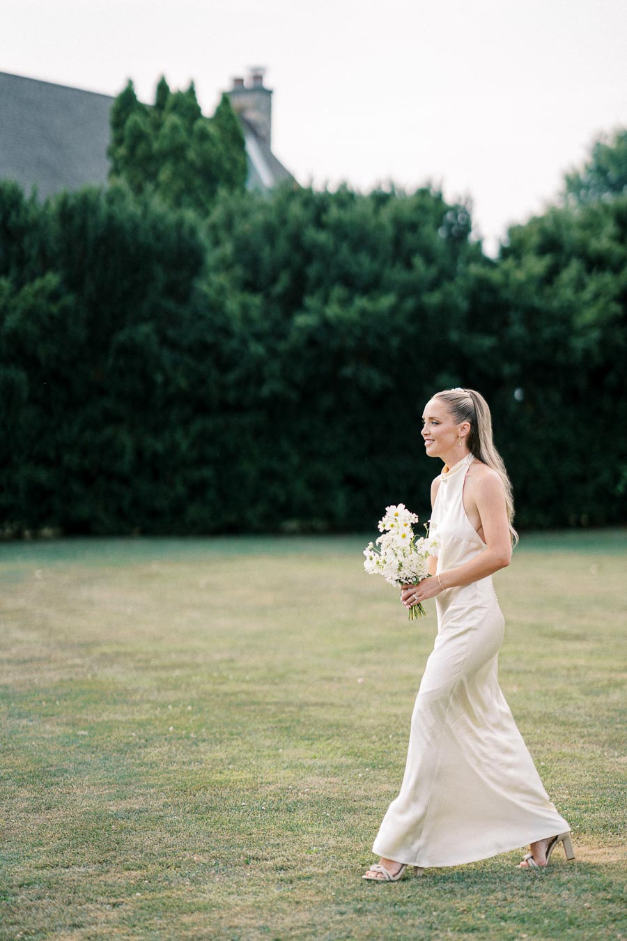 A woman in an elegant ivory dress walking on a grassy lawn, holding a bouquet of white flowers, with lush greenery and a house in the background.