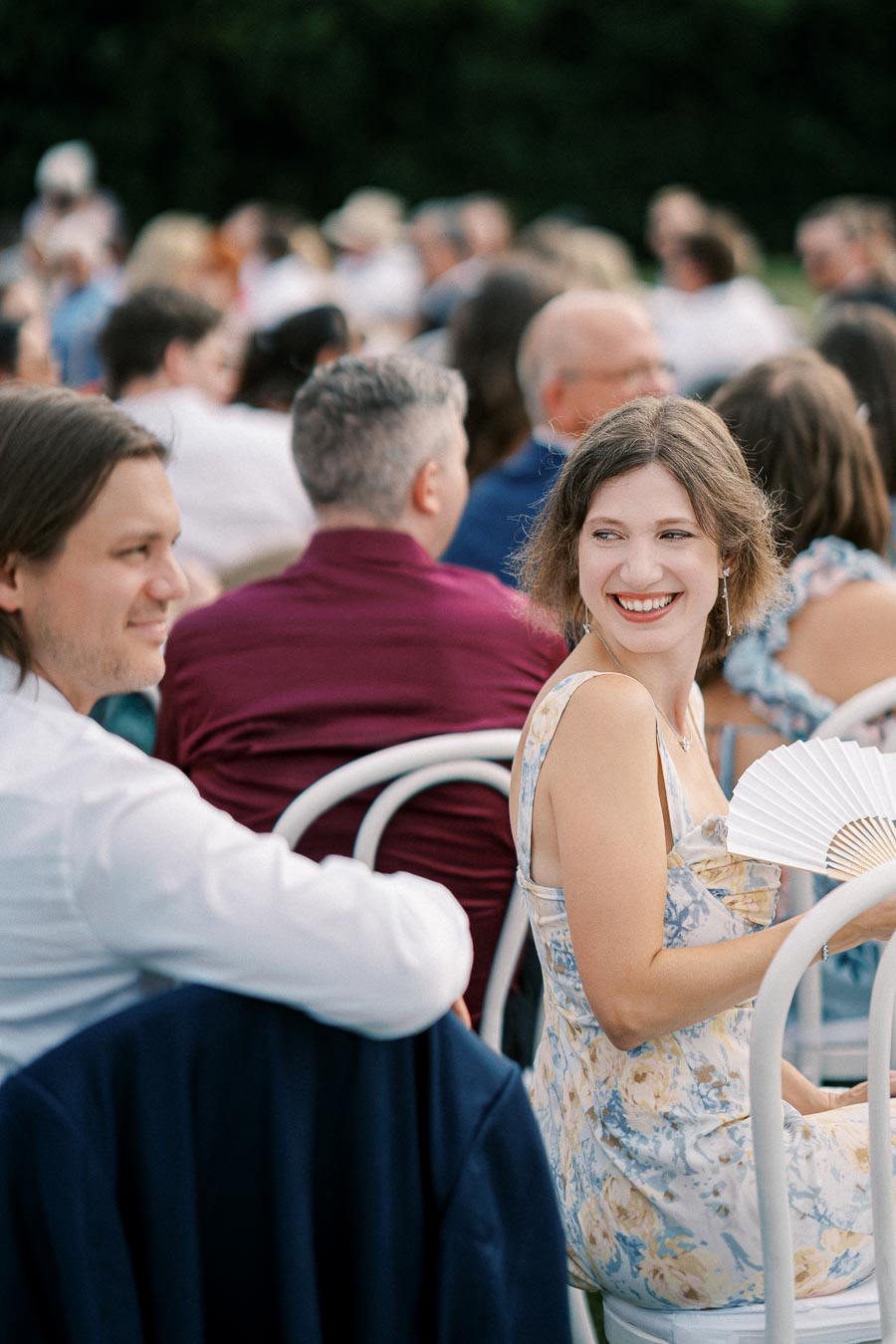 Smiling woman in a summer dress holding a paper fan, sitting outdoors among a group of people at an event.