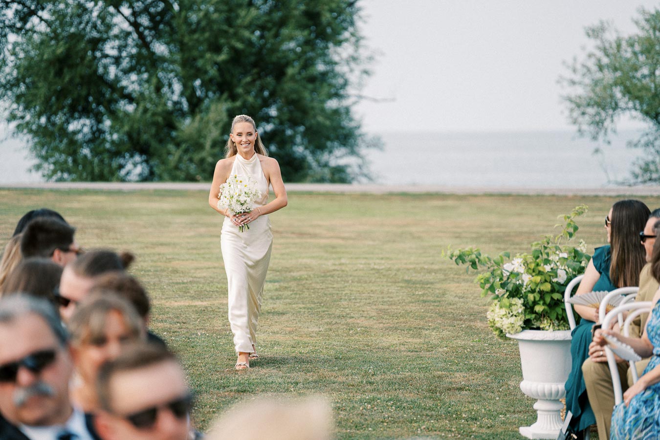 A bride walking down an outdoor aisle in a lush garden setting, holding a bouquet of white flowers, with seated guests in the foreground and a serene lake background.