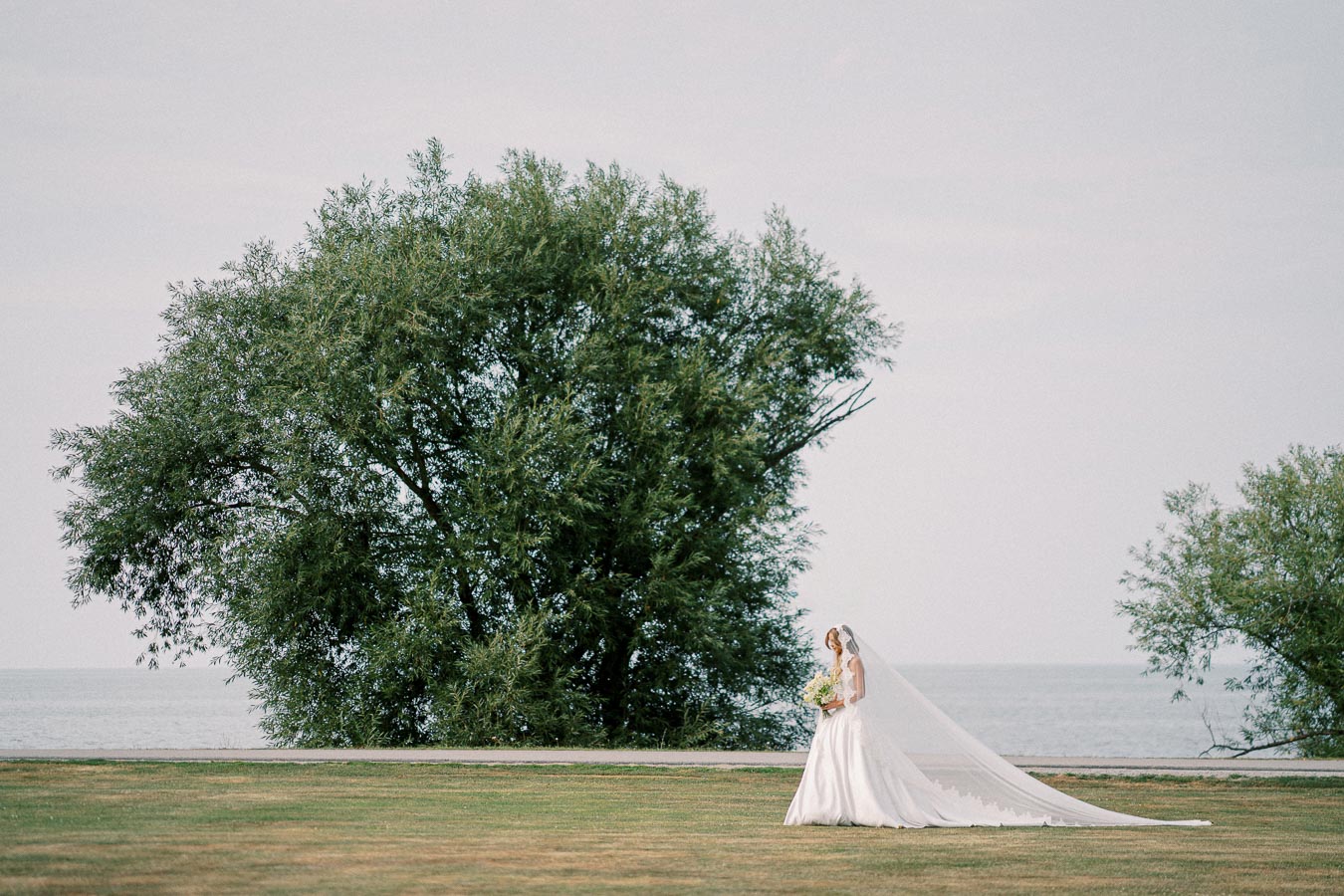 Bride in elegant white gown with long veil holding bouquet, standing near large tree by a serene lake, captured on a clear day.
