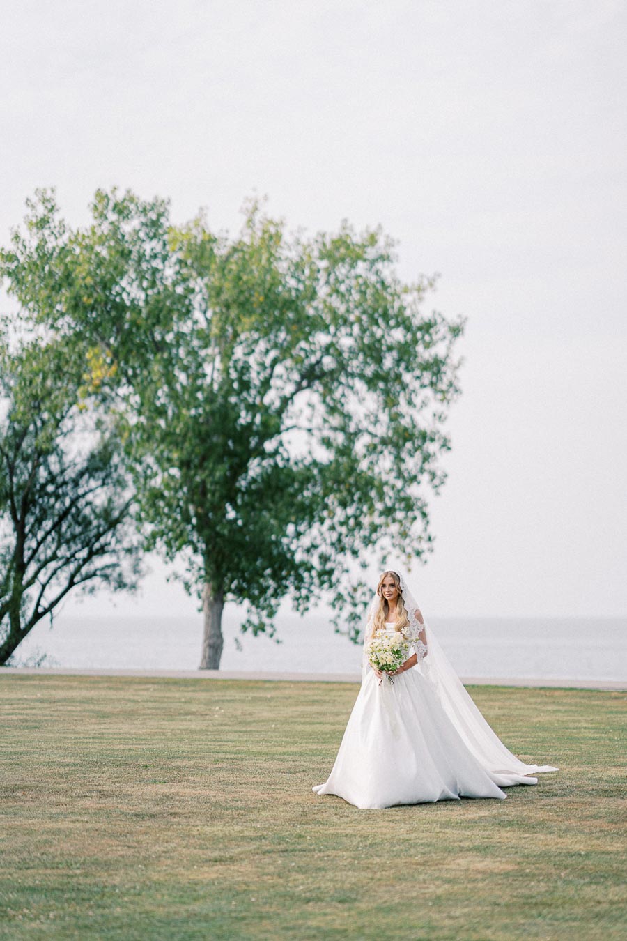 A bride in a flowing white gown and veil holding a bouquet, standing on a grassy field by the water with a large tree in the background.