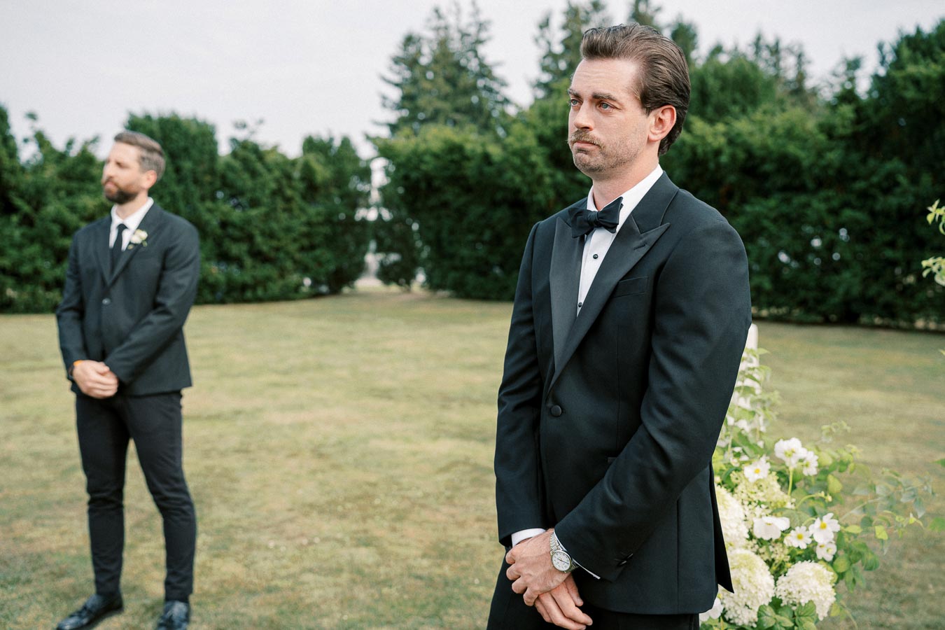 A groom in a black tuxedo looks thoughtfully out into the distance during an outdoor wedding ceremony, standing on green grass with trees in the background, accompanied by the best man also in formal attire.