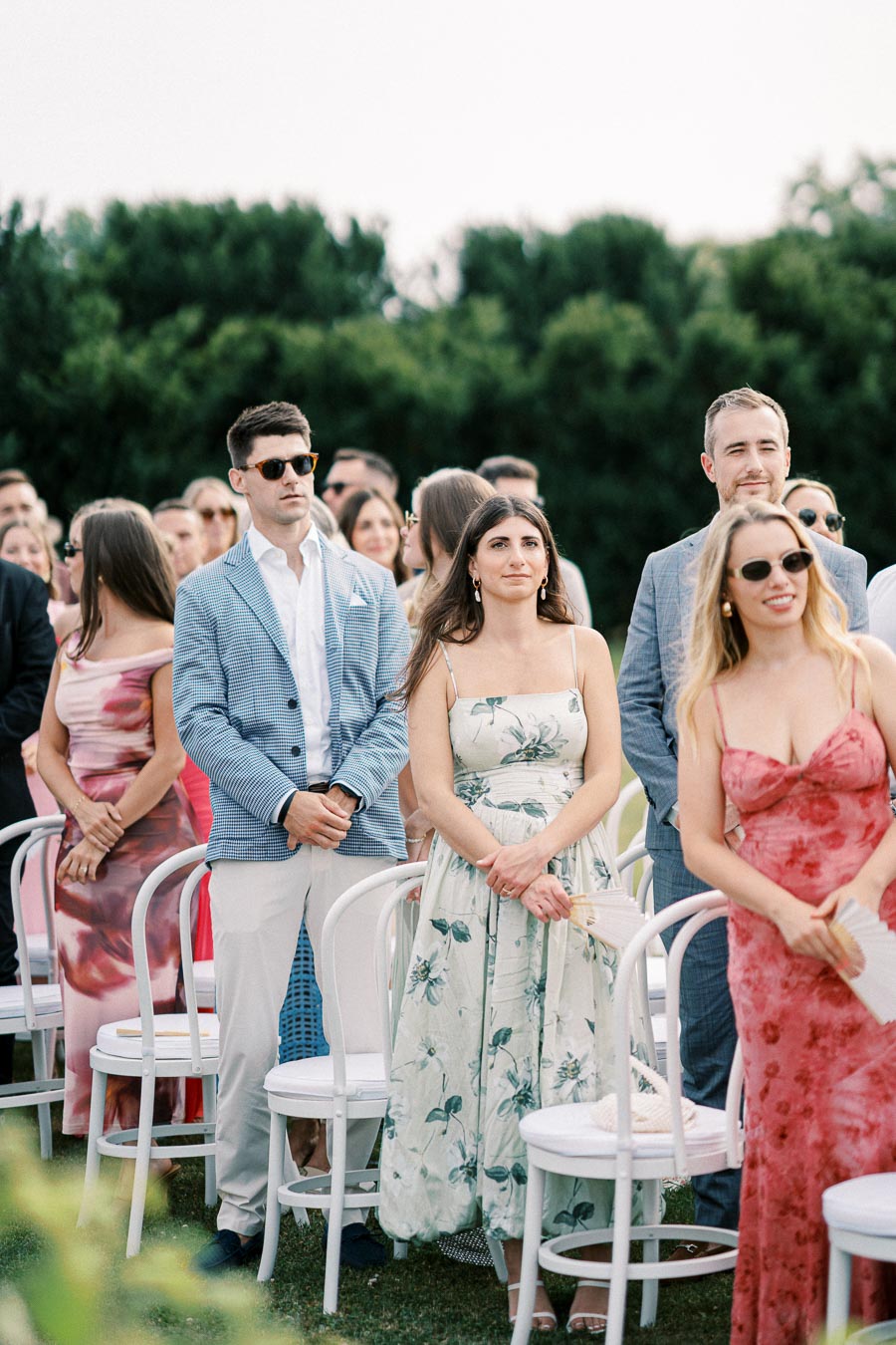 A group of well-dressed guests stands outdoors at a wedding ceremony, with men in suits and women in floral dresses, looking towards the front where the event is happening, surrounded by greenery and white chairs.