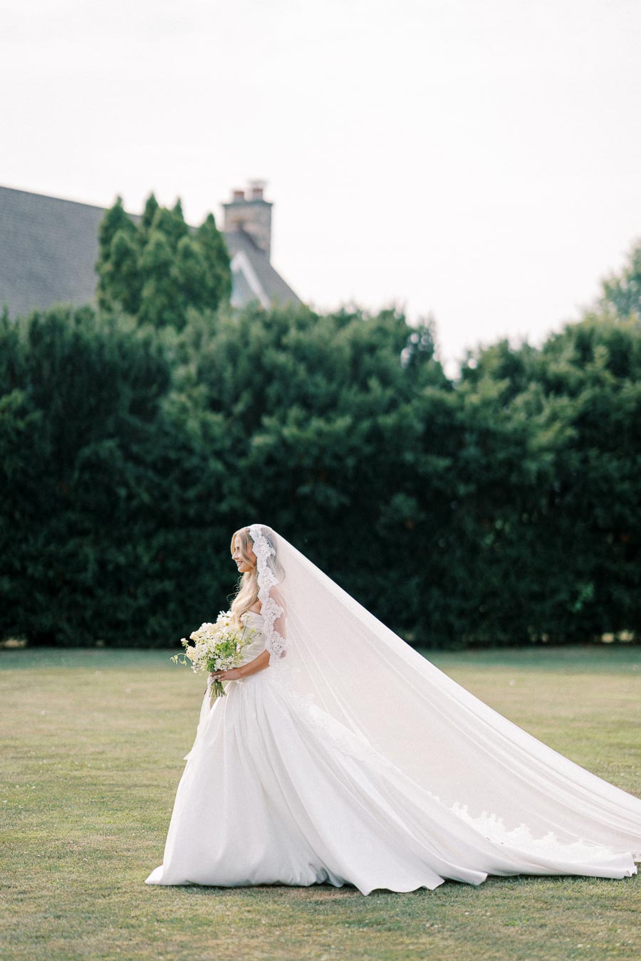 A bride in an elegant white wedding gown with a long lace veil holds a bouquet of flowers while standing outdoors on a grassy lawn, with a house and lush green bushes in the background.