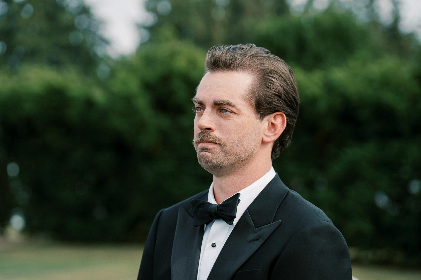 A man in a black tuxedo with a bow tie standing outdoors, with a blurred green background.