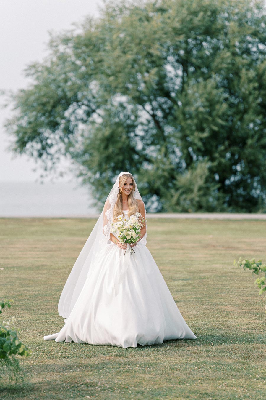 A bride in a white gown and lace veil holding a bouquet of flowers stands on a grassy field with a tree in the background.