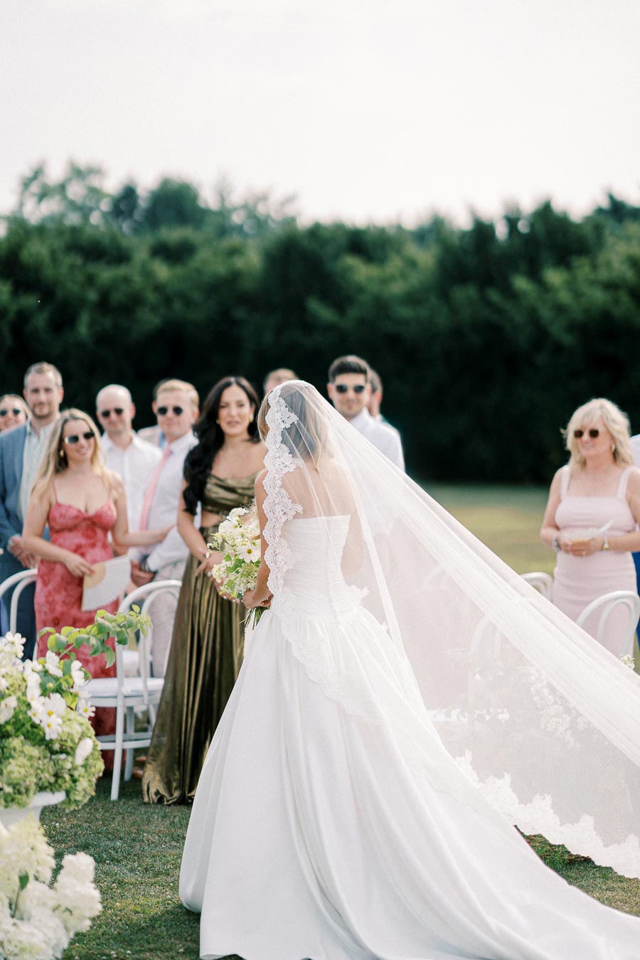 A bride in an elegant white gown walks down the aisle outdoors, surrounded by friends and family, with greenery in the background and vibrant flower arrangements.