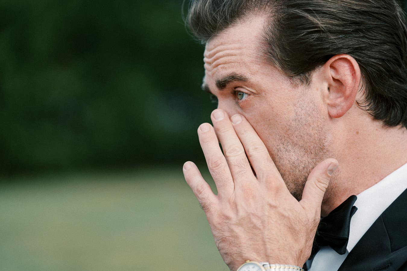 A man in a suit wiping tears from his eye, wearing a watch, with a blurred green background.