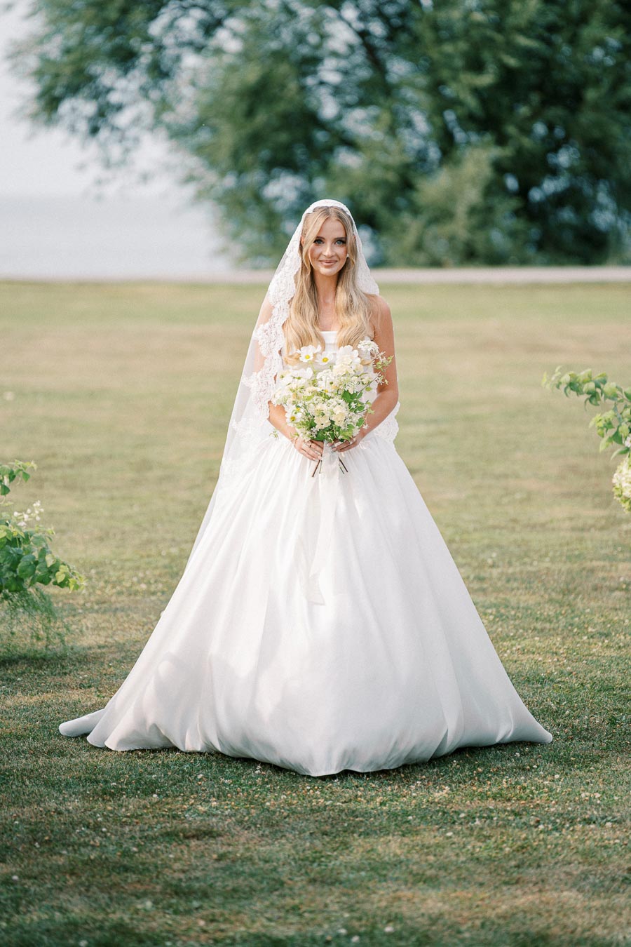 A bride in an elegant white wedding dress stands on a grassy field, holding a bouquet of white and yellow flowers, with lush green trees in the background.