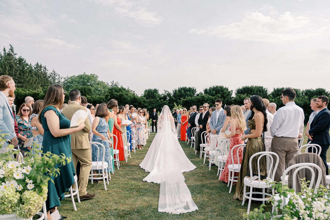 Elegant outdoor wedding ceremony with bride walking down the aisle, surrounded by seated guests in colorful attire under clear skies.