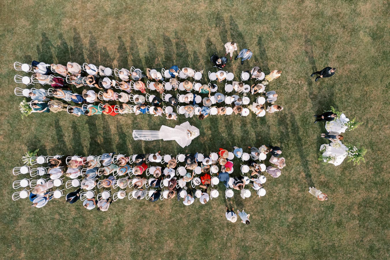 Aerial view of a picturesque outdoor wedding ceremony showing guests seated in rows on a grassy lawn, with a bride and groom at the altar.