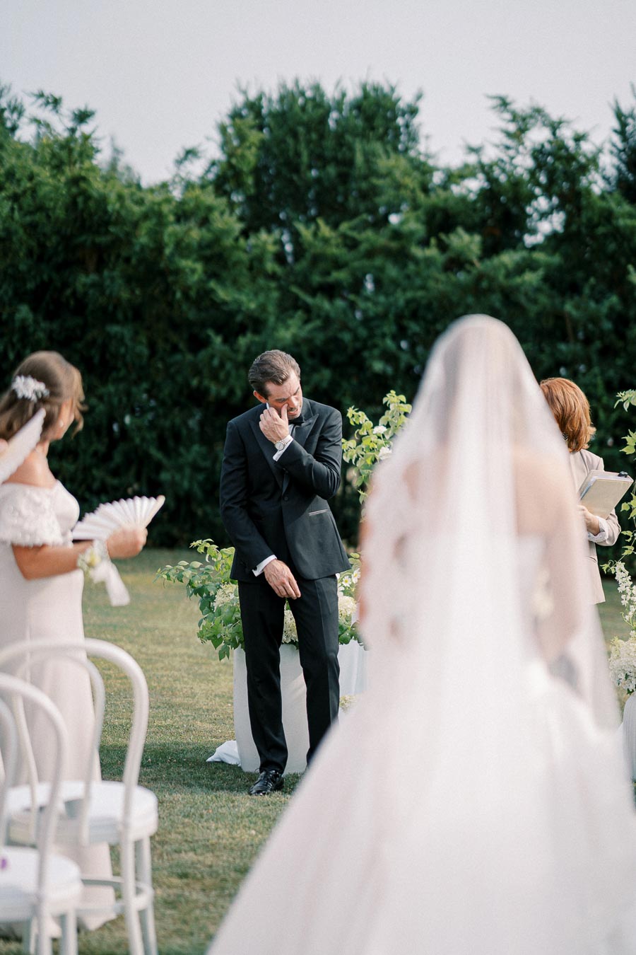 Emotional groom wiping tears during outdoor wedding ceremony, bride in veil approaching, with green foliage backdrop.