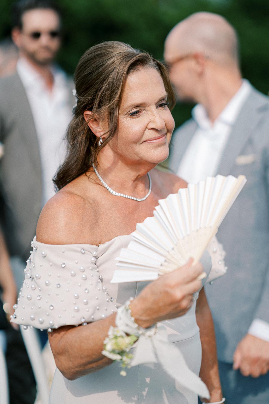 A woman in an elegant, off-shoulder dress adorned with pearls holding a decorative fan at an outdoor event, with people in the background wearing formal attire.
