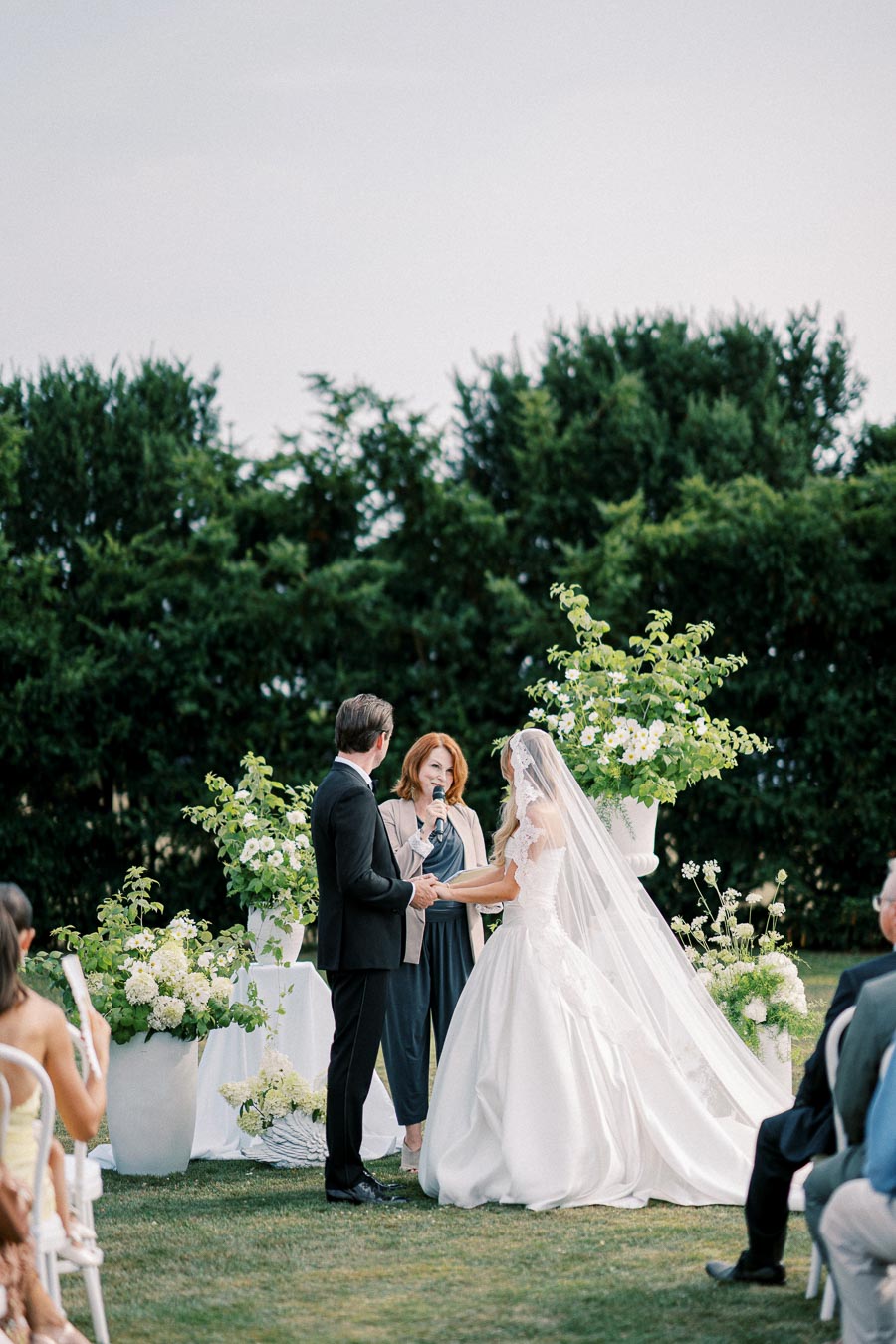 Outdoor wedding ceremony with bride in elegant white gown and groom in black suit exchanging vows, officiated by a woman holding a microphone; surrounded by lush greenery and floral arrangements.