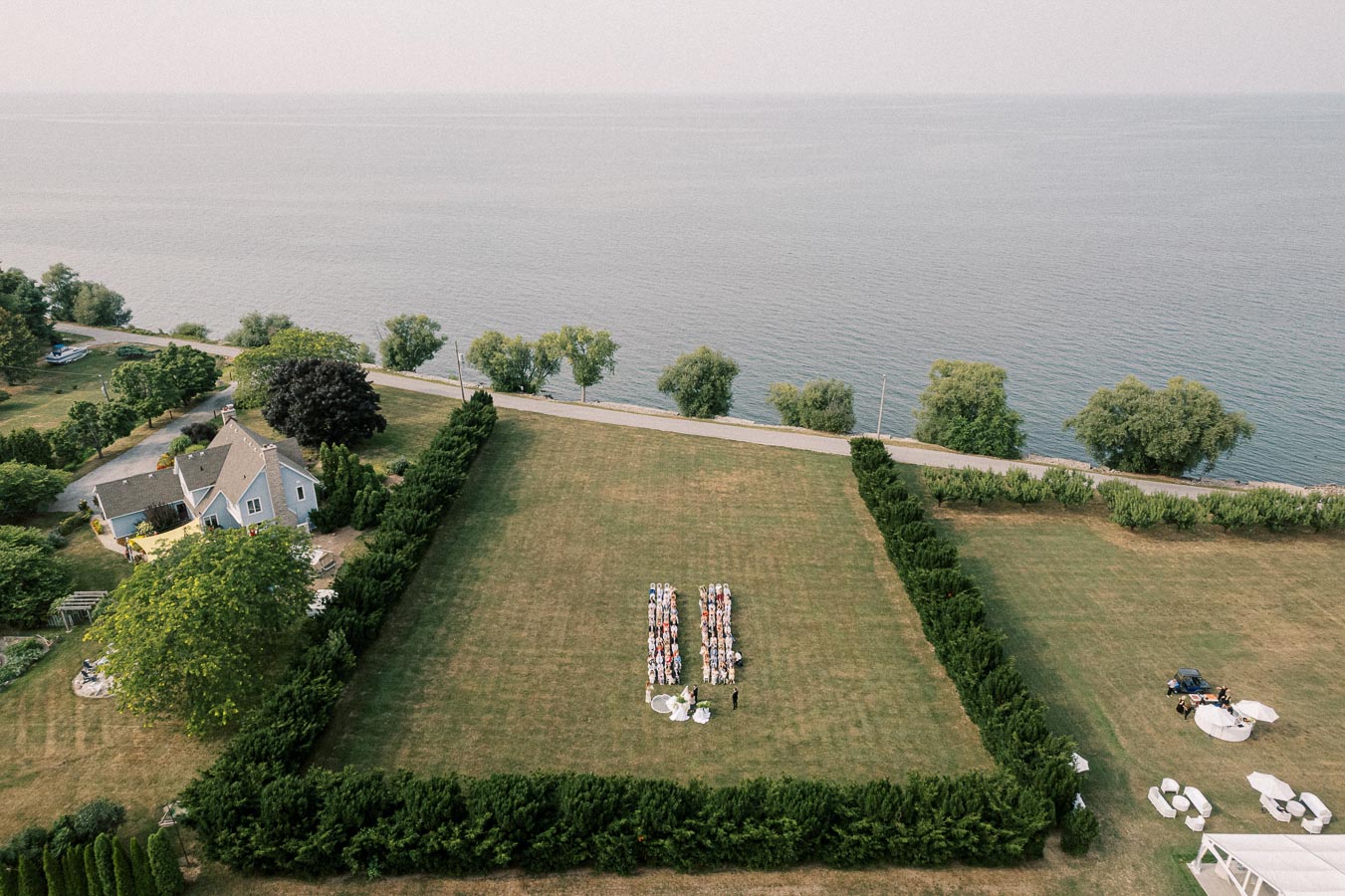 Aerial view of a lakeside wedding ceremony setup with guests seated on a large, manicured lawn bordered by hedges, next to a charming house and a serene body of water in the background.