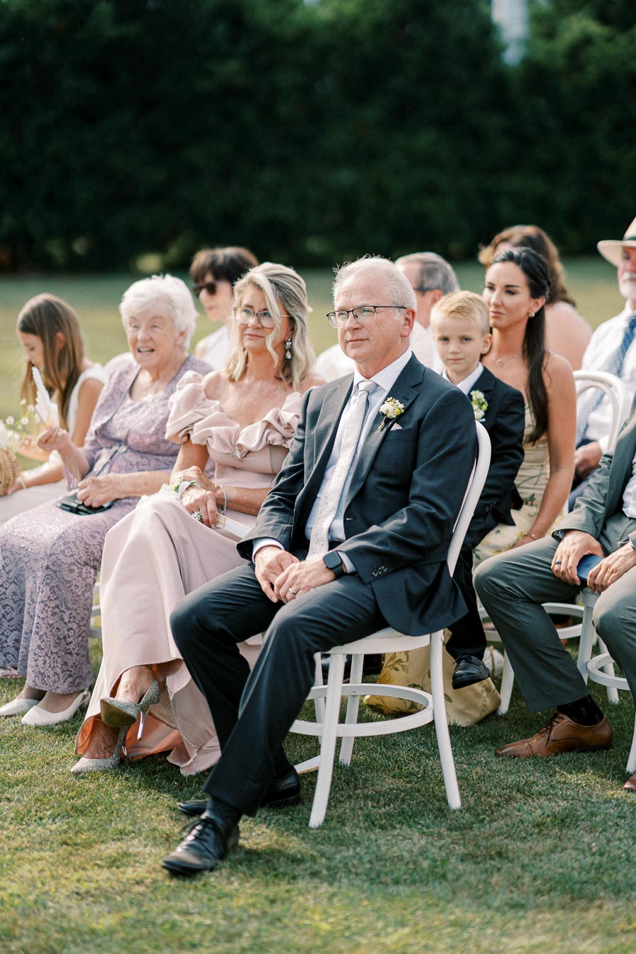 Outdoor wedding ceremony with well-dressed guests seated on white chairs, attentively watching the event.