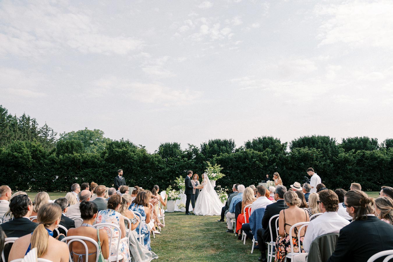 Outdoor wedding ceremony with guests seated on white chairs, a couple exchanging vows in front of lush greenery under a clear sky.