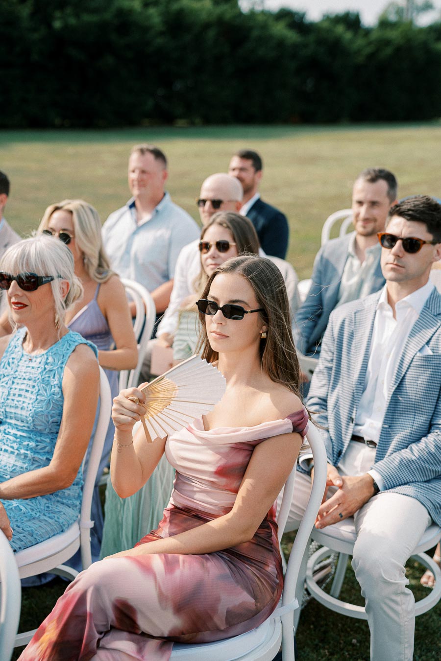 Outdoor wedding ceremony with guests seated, including women in elegant dresses and sunglasses, and a woman fanning herself.