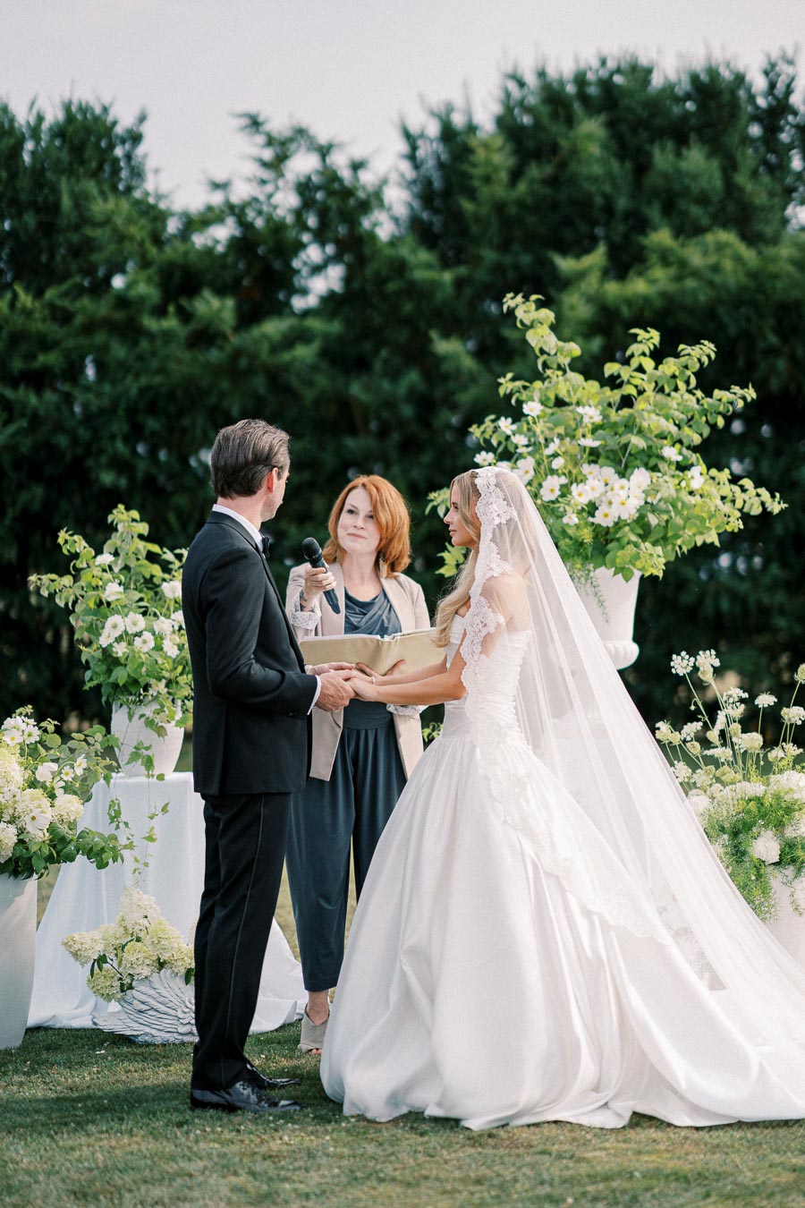 A bride and groom hold hands during an outdoor wedding ceremony, with a celebrant standing beside them. The bride wears a flowing white gown with a lace veil, while the groom is dressed in a black suit. Lush greenery and white floral arrangements adorn the setting, creating an elegant and romantic atmosphere.