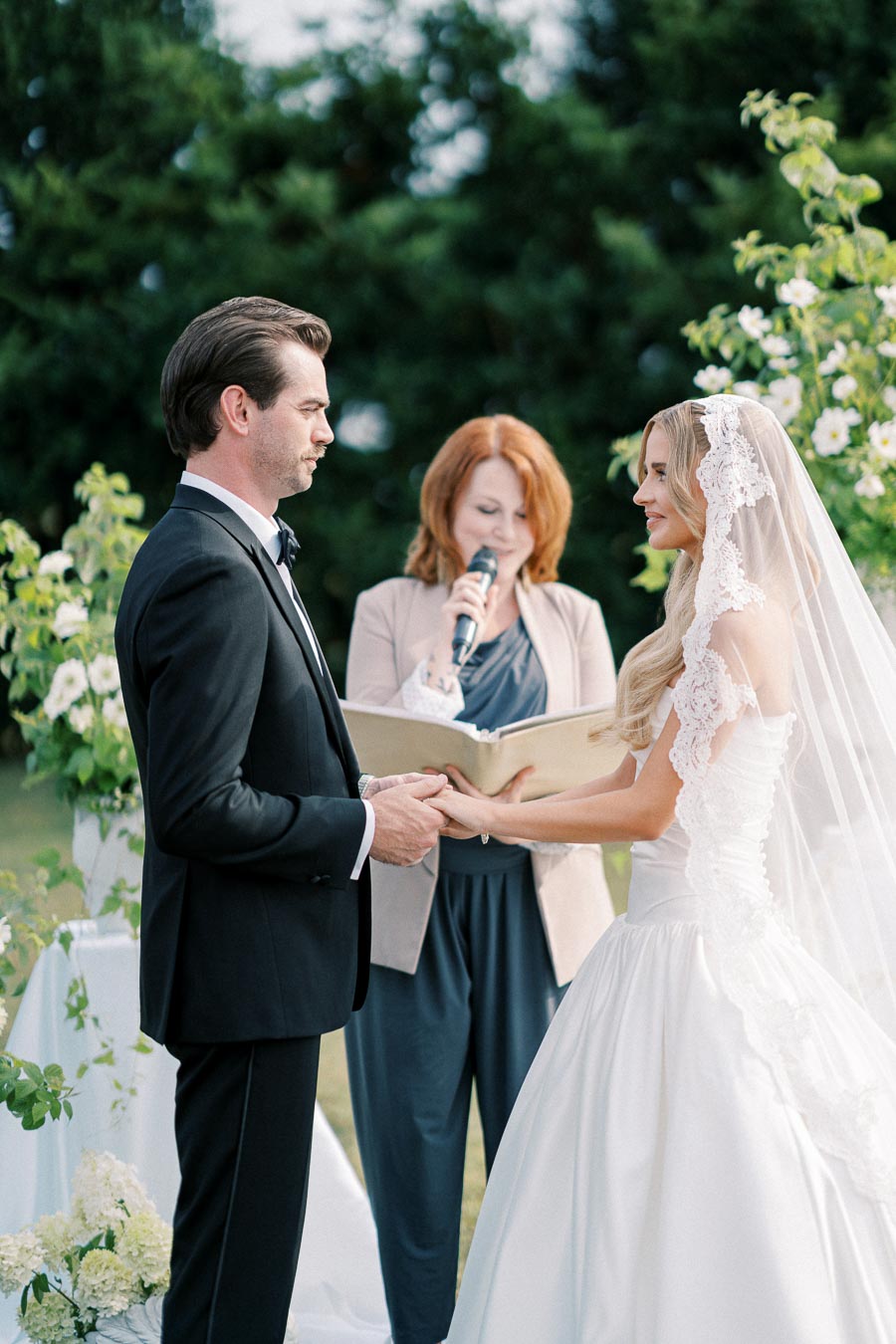 A bride and groom holding hands during an outdoor wedding ceremony, with a female officiant reading vows, surrounded by greenery and floral decorations.