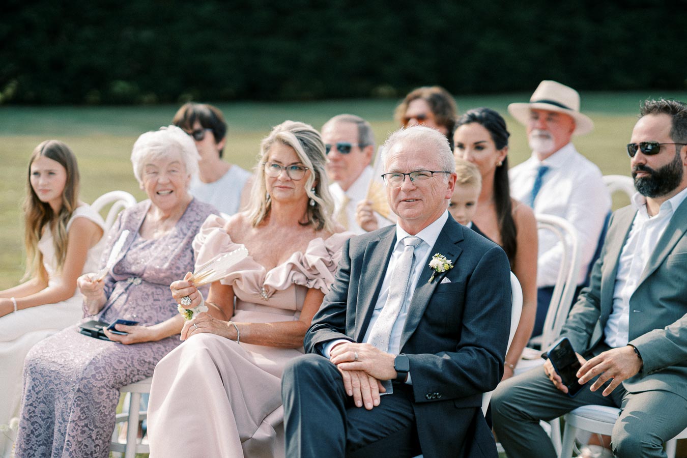 Wedding guests seated and smiling, dressed in formal attire at an outdoor ceremony.