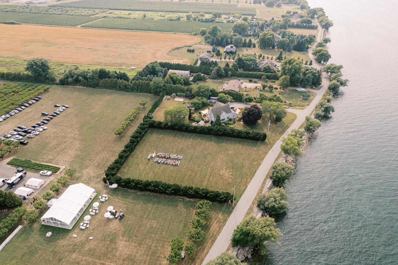 Aerial view of a scenic countryside venue by a waterfront, featuring a large grassy area with arranged seating for an outdoor event, several buildings, parked cars, and lush greenery surrounding the property.