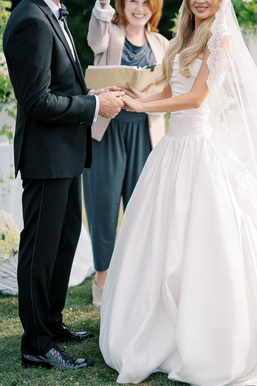 Bride and groom exchanging vows outdoors with officiant smiling, showcasing elegant wedding ceremony.