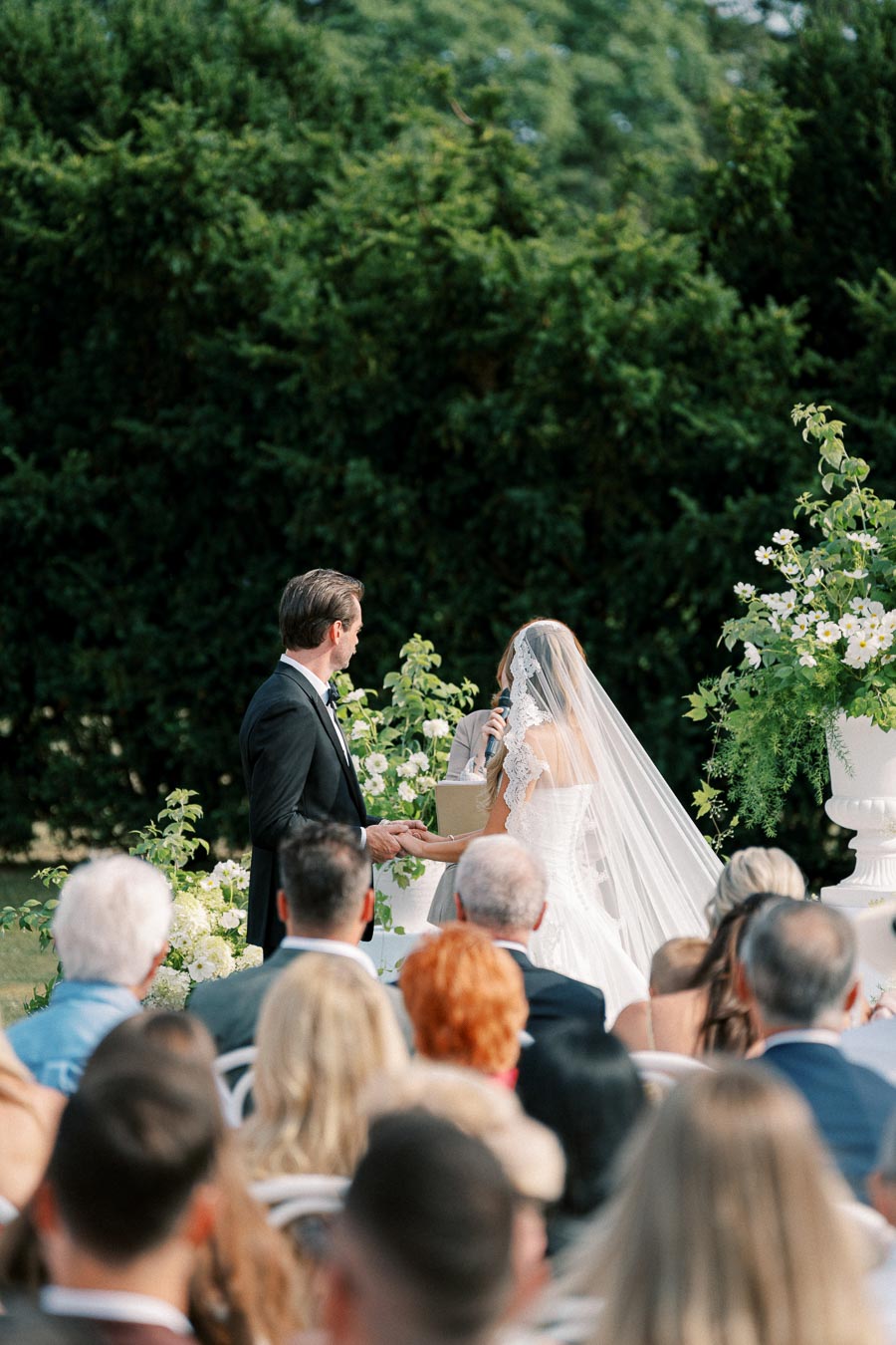 Outdoor wedding ceremony with a couple exchanging vows, surrounded by guests and lush greenery. Bride in a white gown with a veil, groom in a black suit.