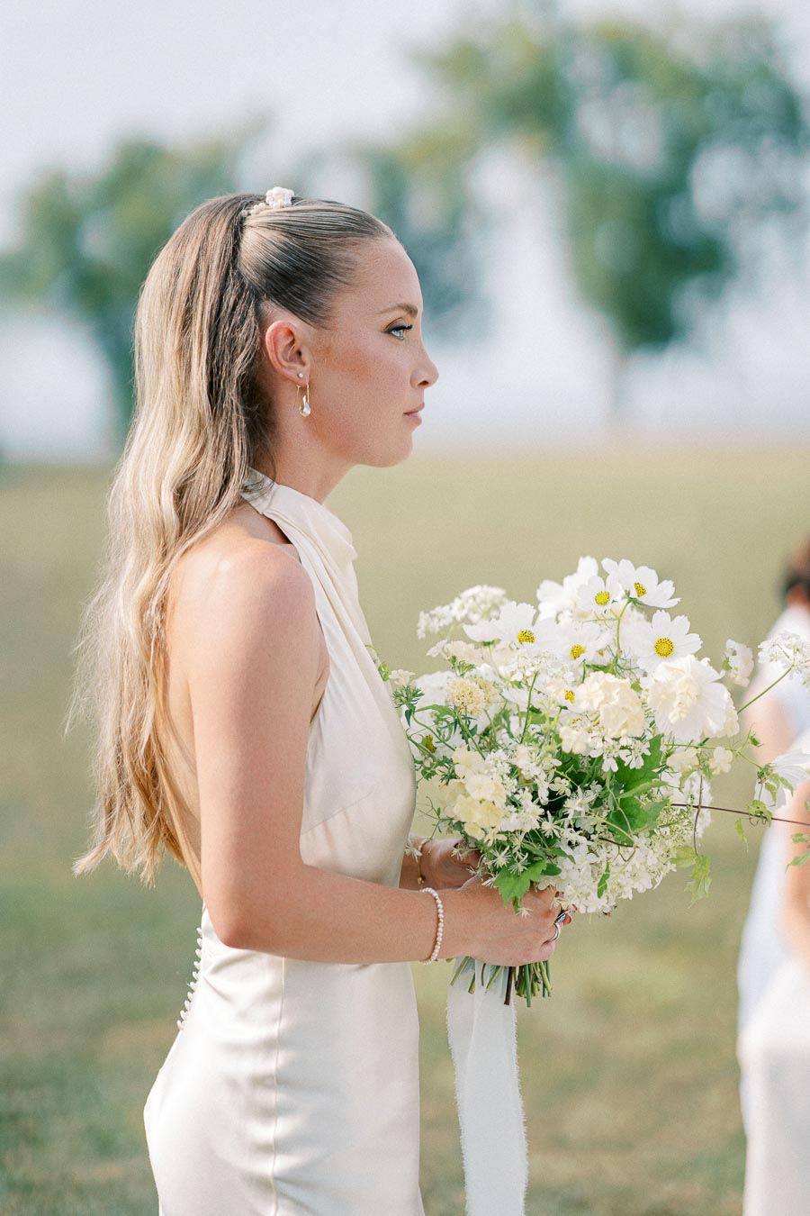 A woman in a cream-colored dress holds a bouquet of white wildflowers outdoors, with a blurred natural background featuring trees and a grassy field.
