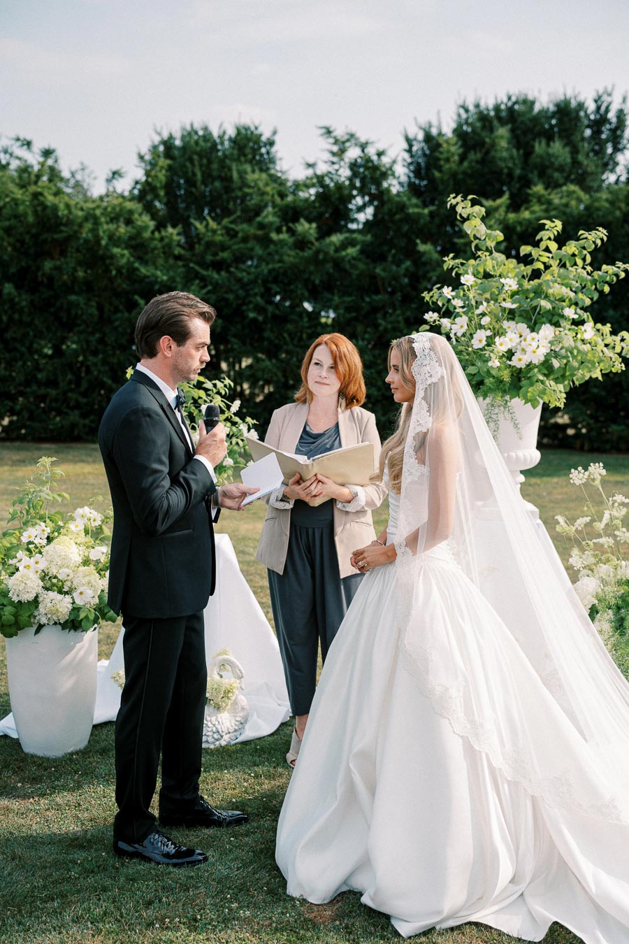 Outdoor wedding ceremony with a bride in a lace wedding gown, groom in a black suit, and officiant holding a book, surrounded by white floral arrangements on green lawn.