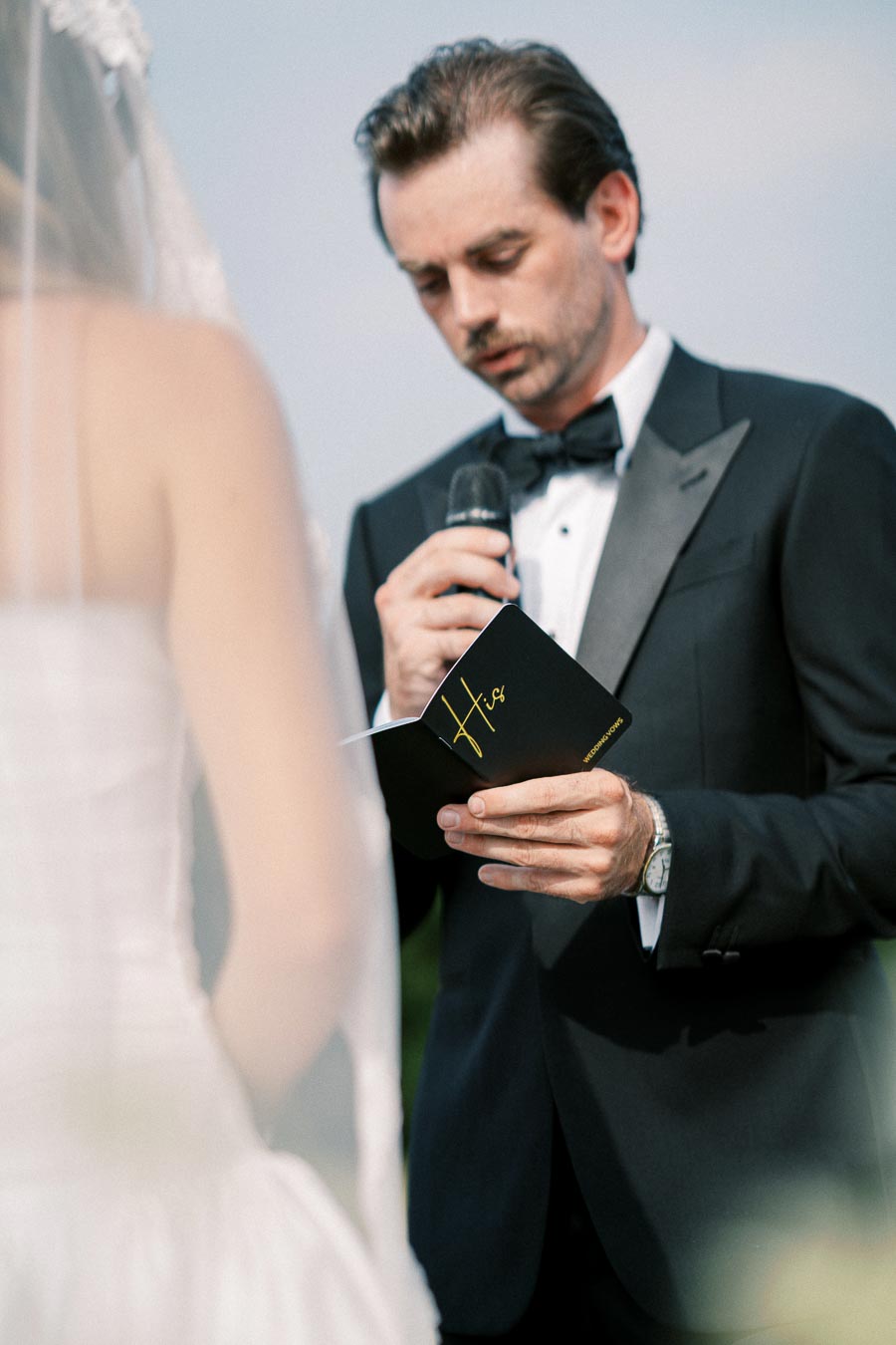 Groom in a tuxedo reading vows from a black booklet during an outdoor wedding ceremony.