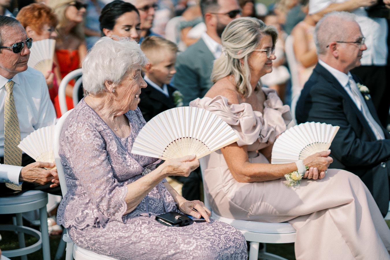 Elderly guests seated at an outdoor event, holding hand fans to keep cool in the warm weather.
