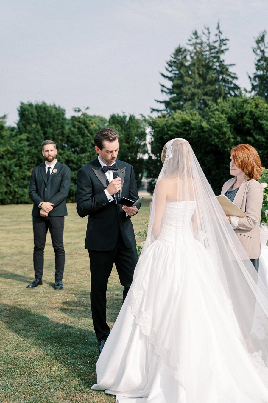 A groom in a black tuxedo reads vows to his bride, wearing a white wedding gown and veil, during an outdoor wedding ceremony, with an officiant and a groomsman in the background under a clear sky.