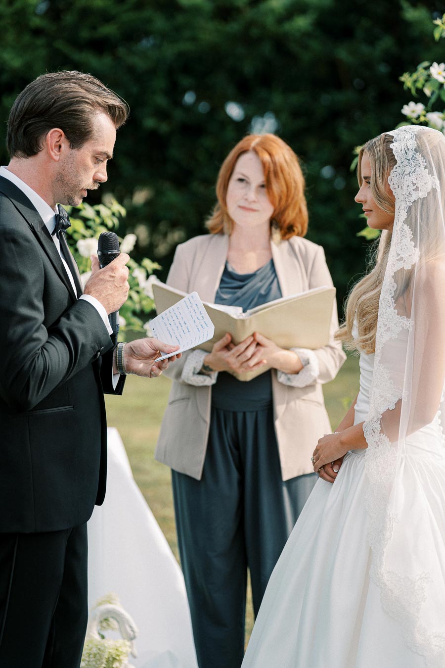 A groom in a black suit reads vows from a small notebook, standing beside the bride in a white wedding dress with a lace veil, while an officiant looks on, outdoors with greenery in the background.