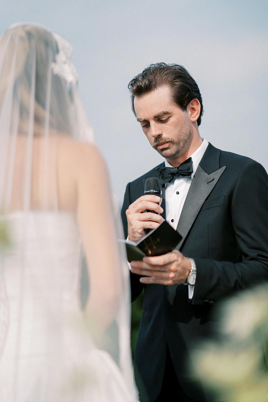 Groom in a black tuxedo reading vows to bride in a wedding ceremony, holding a microphone and book, with a clear sky background.