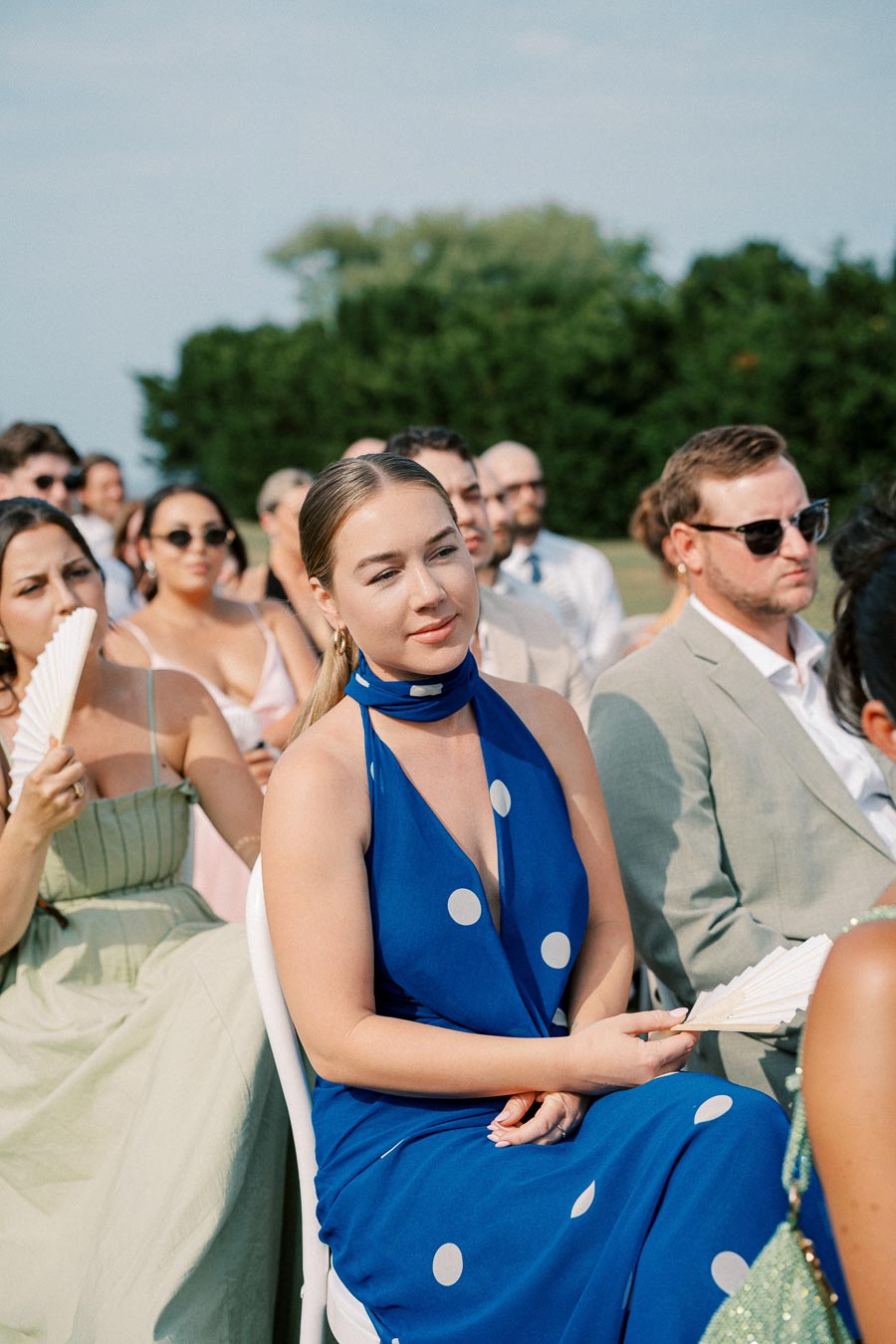 Wedding guests seated outdoors, woman in blue polka dot dress and others using hand fans, under a clear sky with greenery in the background.
