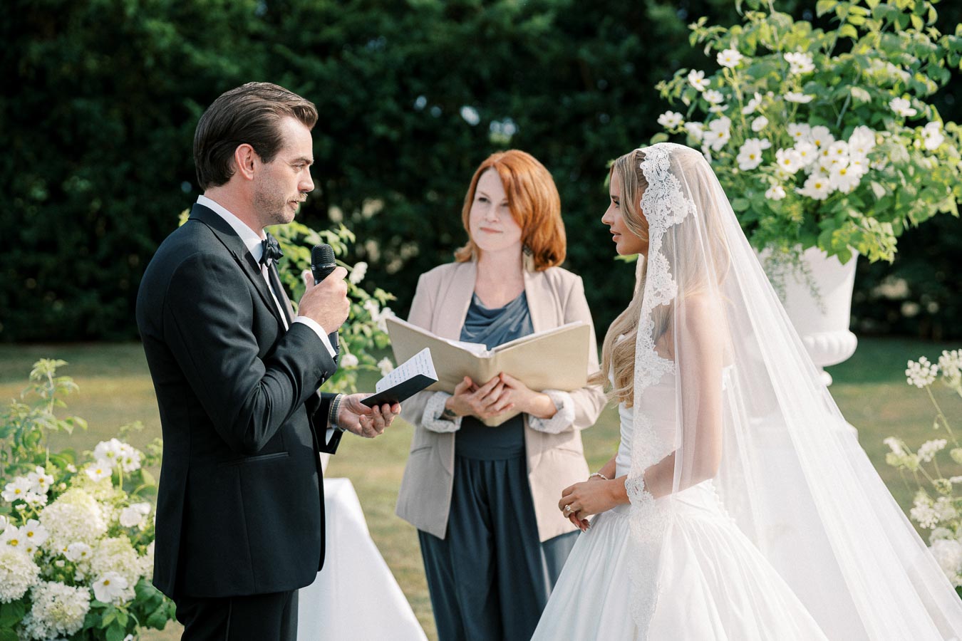 Groom and bride exchanging vows during outdoor wedding ceremony, with officiant standing between them, surrounded by lush greenery and white flowers.