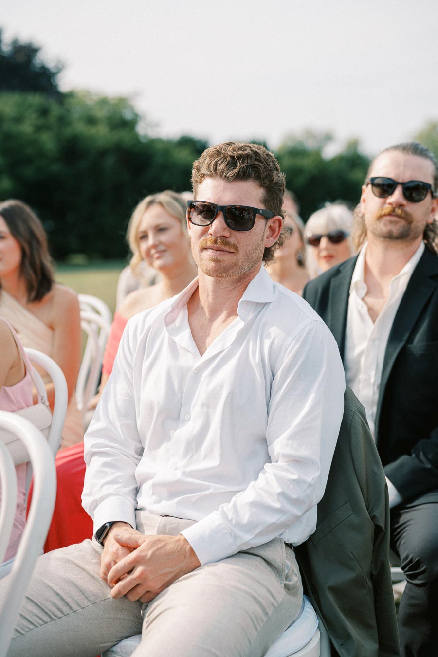 A man in a white shirt and sunglasses sits attentively at an outdoor event, surrounded by other guests. The setting appears formal, with individuals dressed in elegant attire, suggesting a wedding or ceremony. Greenery in the background adds to the serene atmosphere.