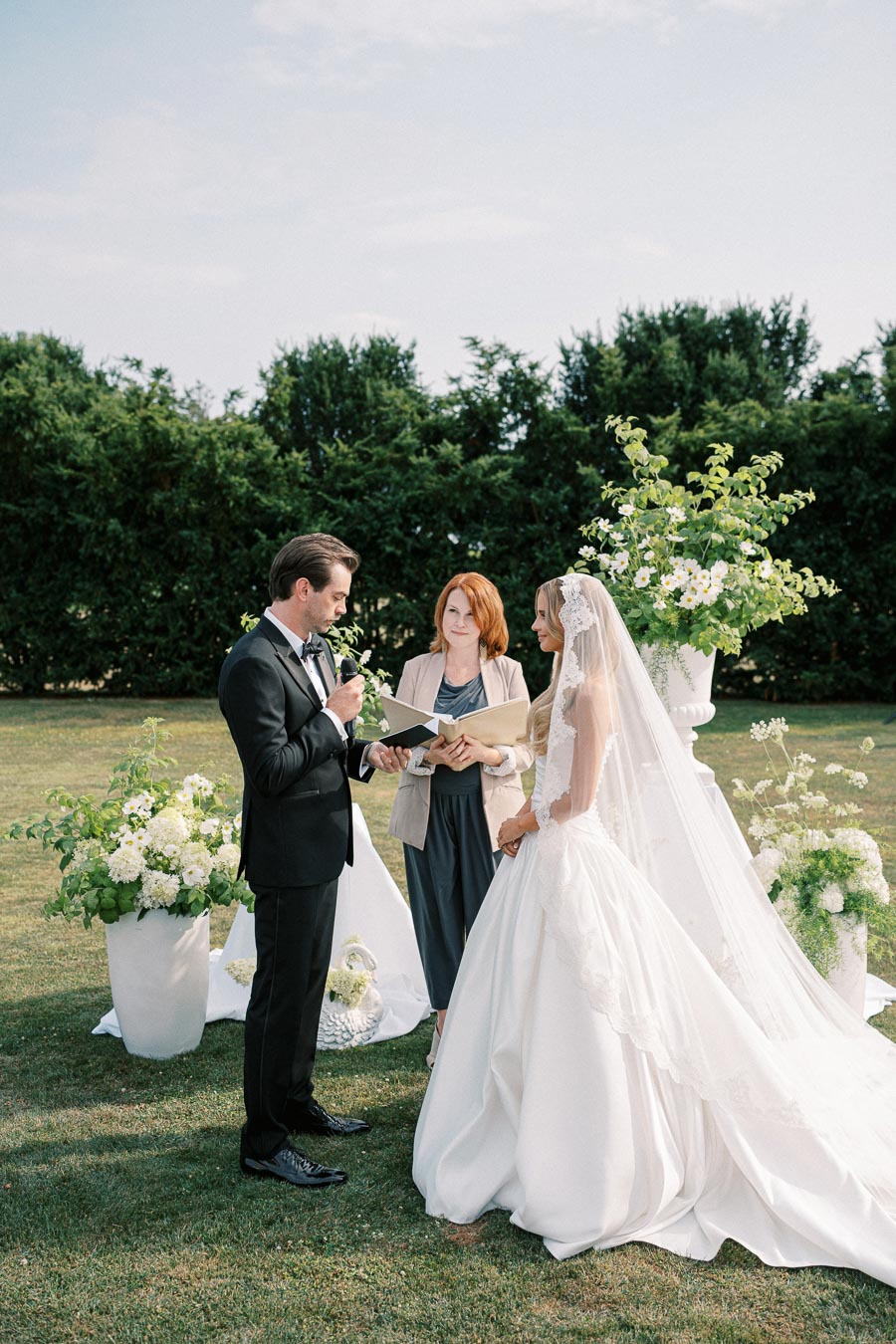 Outdoor wedding ceremony with couple exchanging vows, officiant present, surrounded by lush greenery and floral arrangements.