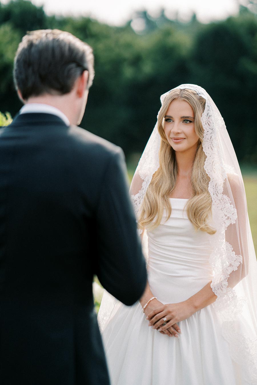A bride in a white wedding dress with a lace veil stands outdoors, facing a groom in a black suit during a wedding ceremony.